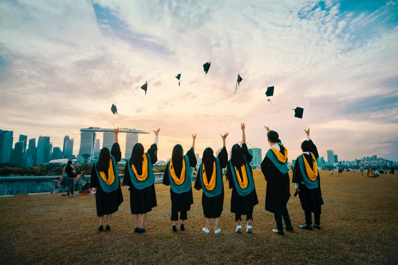 Graduates in gowns seen from behind throwing caps into the air at dusk.